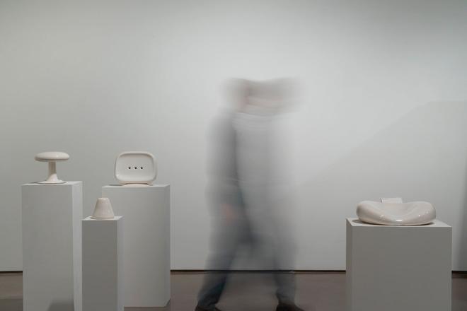 A person walking pass collection of white ceramic objects installed on white pedestals 