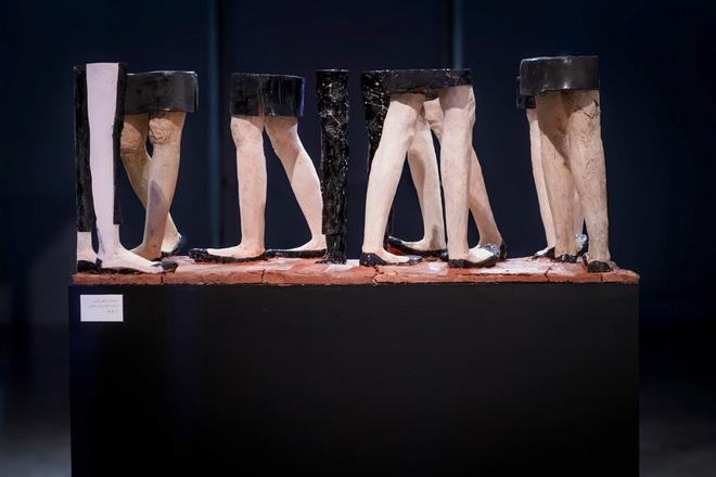 Sculpture of eight pairs of human legs from the thighs down, two wearing trousers with the other six wearing skirts, 展示在一个黑暗的平台上，上面有一层红色的粘土, 在画廊设置中.